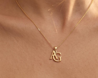 May include: A gold-toned necklace featuring a pendant with the interlocking capital letters A and G. The pendant hangs from a delicate, thin gold-toned chain that rests against the collarbone. The letters are rendered in a classic serif font.