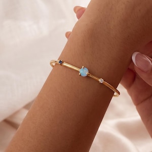 May include: A gold-toned bangle bracelet featuring a central light blue round gemstone, flanked by a smaller dark blue stone on one side and a clear white stone on the other, set along a thin, polished metallic band.