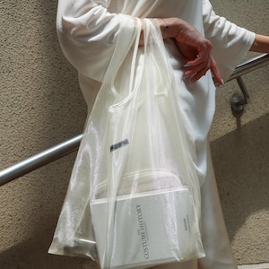 Könnte beinhalten: Eine durchscheinende, cremeweiße Organza-Tragetasche mit zwei dünnen Henkeln. In der Tasche befindet sich ein weißes Hardcover-Buch mit dem Titel The Costume History von Taschen und schwarzem Text auf dem Einband. Ein kleines, rechteckiges schwarzes Etikett.