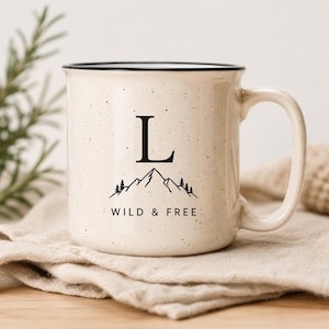 Könnte beinhalten: Ein cremefarbener Keramikbecher mit gesprenkelter Oberfläche und schwarzem Rand. Die Vorderseite zeigt eine schwarze Grafik eines Gebirges mit Tannen, den Buchstaben L über den Bergen und den Text WILD & FREE darunter gedruckt.