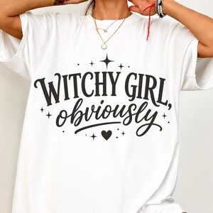 May include: A white short-sleeved t-shirt featuring black text that reads "WITCHY GIRL, obviously" in a mix of serif and script fonts. The design is accented with small black stars and a heart symbol centered below the text.
