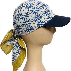 May include: A head covering featuring a white fabric base with a blue floral pattern and a solid dark navy blue brim. The back of the head covering includes two fabric ties that hang down, displaying a mustard yellow border on the underside.