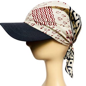 May include: A head covering featuring a black peak and a patterned fabric top. The fabric is cream-coloured with a repeating pattern of red zigzags and small floral shapes. The back has a long, patterned tail.