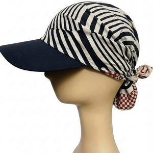 May include: A cap featuring a solid dark navy blue brim and a crown made of cream fabric with black diagonal stripes. The back of the cap includes a tied fabric section with a small red and white checkered pattern and a geometric print design.
