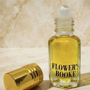 May include: A clear glass roll-on bottle filled with a golden yellow liquid, featuring a black label that reads FLOWER'S BOOKE. The bottle has a transparent roller ball top and is accompanied by a separate, shiny gold-colored cylindrical cap.