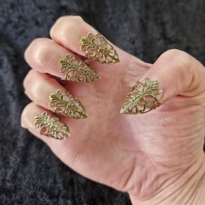 Könnte beinhalten: Fünf antike, goldfarbene Nagelringe, jeweils mit einem verzierten, durchbrochenen Filigranmuster. Die Ringe sind so geformt, dass sie die Fingernägel bedecken und eine dramatische und gotische Ästhetik erzeugen. Die Ringe werden auf einer Hand präsentiert.