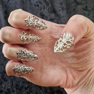 Könnte beinhalten: Silberfarbene Nagelringe mit einem verzierten Filigran-Design. Die Ringe bedecken die Fingernägel und sind so geformt, dass sie spitzen Krallen ähneln. Die Hand wird vor einem dunklen Hintergrund gezeigt, der die filigrane Metallarbeit hervorhebt.