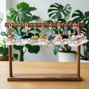 May include: A wooden display stand holds ten small, crocheted ghost keychains. Each ghost features a white body with pink cheeks and wears a unique, colorful crocheted hat, including designs like a pumpkin, animal ears, and various patterned caps.
