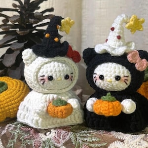 Puede incluir: Dos figuras de gato tejidas a ganchillo con sombreros de bruja y pequeñas calabazas naranjas. Una figura es blanca con sombrero negro y lazo rojo, la otra es negra con sombrero blanco y lazo rosa. Ambas tienen estrellas amarillas en sus sombreros.