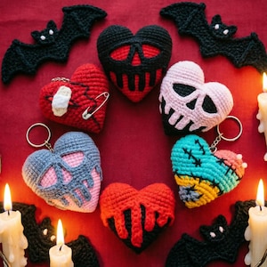May include: A selection of handmade crochet heart-shaped keyrings in various colours and designs, including skull and stitched patterns. Black crochet bats and lit candles surround the hearts on a red background, creating a spooky, Halloween-themed display.