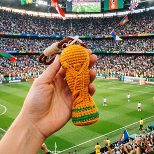 May include: A yellow and green crocheted replica of the FIFA World Cup trophy keychain. The item features a textured, yarn-based construction with a brown cord loop attached to the top, mimicking the iconic shape of the global soccer tournament award.