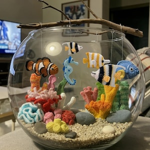 Könnte beinhalten: Eine klare Glasschale mit einer Sammlung gehäkelter Meeresbewohner, darunter ein Clownfisch, ein Seepferdchen und verschiedene tropische Fische, die an einem Ast hängen. Der Boden ist mit kleinen Kieselsteinen, glatten Steinen und gehäkelten Korallen gefüllt.