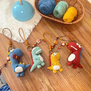 Pode incluir: Quatro porta-chaves de dinossauro em croché nas cores azul, verde claro, amarelo e vermelho. Cada um apresenta uma barriga branca, dentes brancos e uma alça tipo couro com pequenas contas coloridas e botões presos a um anel de metal.