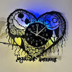 Può includere: Un orologio da parete nero a forma di cuore con Jack e Sally da The Nightmare Before Christmas. L'orologio ha un quadrante nero con lancette e numeri bianchi. Le parole "Together Forever" sono in basso e l'orologio è illuminato con luci blu e gialle.