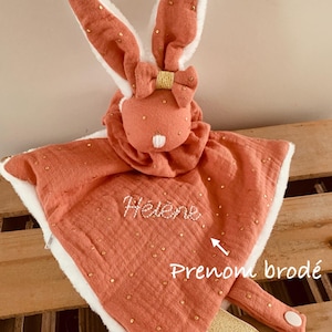 Op de afbeelding: Een koraalkleurige babyknuffel met een konijnenkop en lange oren. Het konijn heeft een strik en is versierd met goudkleurige stippen. De deken heeft een witte, pluizige rand en de naam "Hélène" is erop geborduurd.