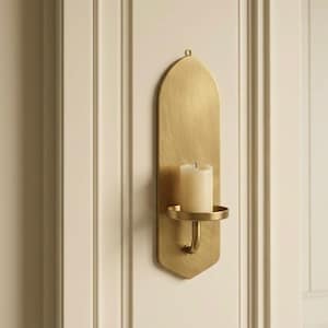 May include: A gold-colored wall sconce with a candle. The sconce has an arched top and a curved arm holding a round candle holder. The candle is white and cylindrical. The sconce is mounted on a cream-colored wall.