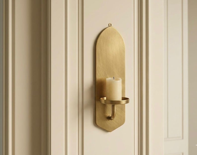 May include: A gold-colored wall sconce with a candle. The sconce has an arched top and a curved arm holding a round candle holder. The candle is white and cylindrical. The sconce is mounted on a cream-colored wall.