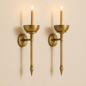 May include: A pair of wall-mounted candle sconces crafted from brushed brass metal. Each sconce features a circular wall plate, a slender vertical arm ending in a pointed finial, and a shallow bowl-shaped candle holder containing a lit taper candle.