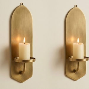 May include: Two brass wall sconces with a warm, golden finish. Each sconce features a long, arched backplate, a circular candle holder, and a lit white candle. The sconces are designed to hold pillar candles and add a touch of elegance.