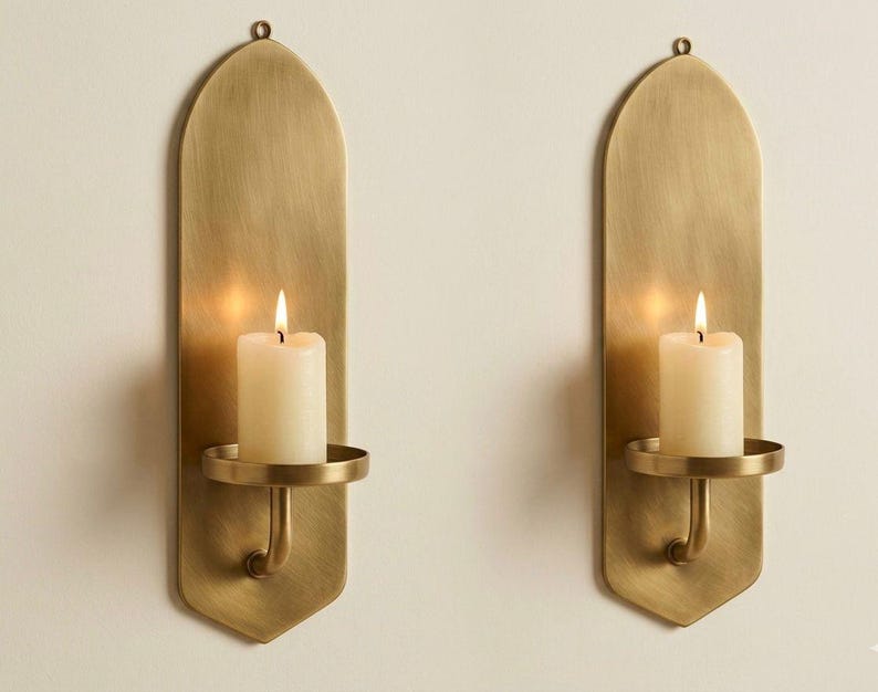 May include: Two brass wall sconces with a warm, golden finish. Each sconce features a long, arched backplate, a circular candle holder, and a lit white candle. The sconces are designed to hold pillar candles and add a touch of elegance.