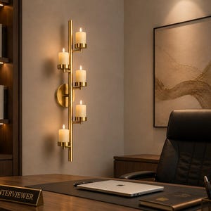 May include: A vertical wall-mounted candle holder in a brushed gold finish featuring five cylindrical candle cups arranged along a central rod, each holding a lit white pillar candle, attached to the wall with a circular gold mounting plate.