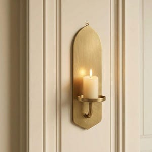 May include: A brass wall sconce with a lit white candle. The sconce has a curved design with a small circular holder for the candle. The sconce is mounted on a white wall.