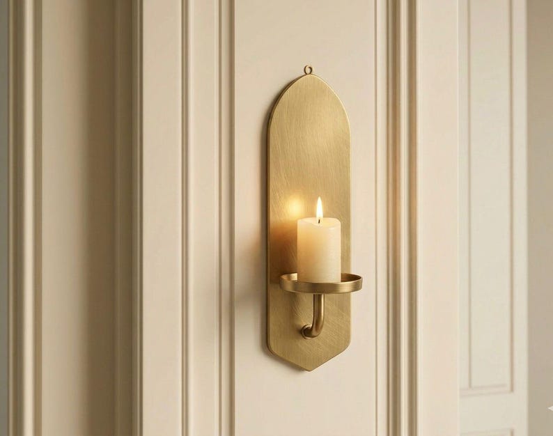 May include: A brass wall sconce with a lit white candle. The sconce has a curved design with a small circular holder for the candle. The sconce is mounted on a white wall.