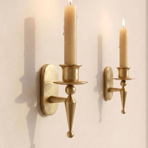 May include: Two wall-mounted candle holders made of brushed brass with a conical base and a circular drip plate. Each holder features a vertical oval backplate and supports a single cream-colored taper candle with a lit flame, casting a shadow.