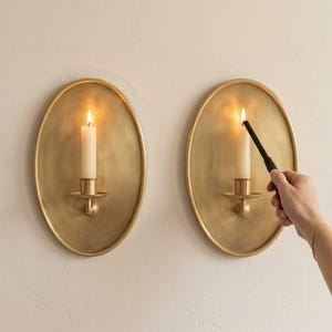May include: Two oval brass wall sconces with a brushed finish, each featuring a central candle holder. One white pillar candle is lit, while a black lighter ignites the wick of the second white pillar candle mounted on the wall.