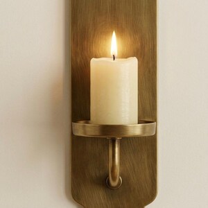 May include: A lit ivory candle sits in a brass wall sconce. The sconce has a rectangular backplate and a circular candle holder. The flame is bright against the warm tones of the brass and candle.