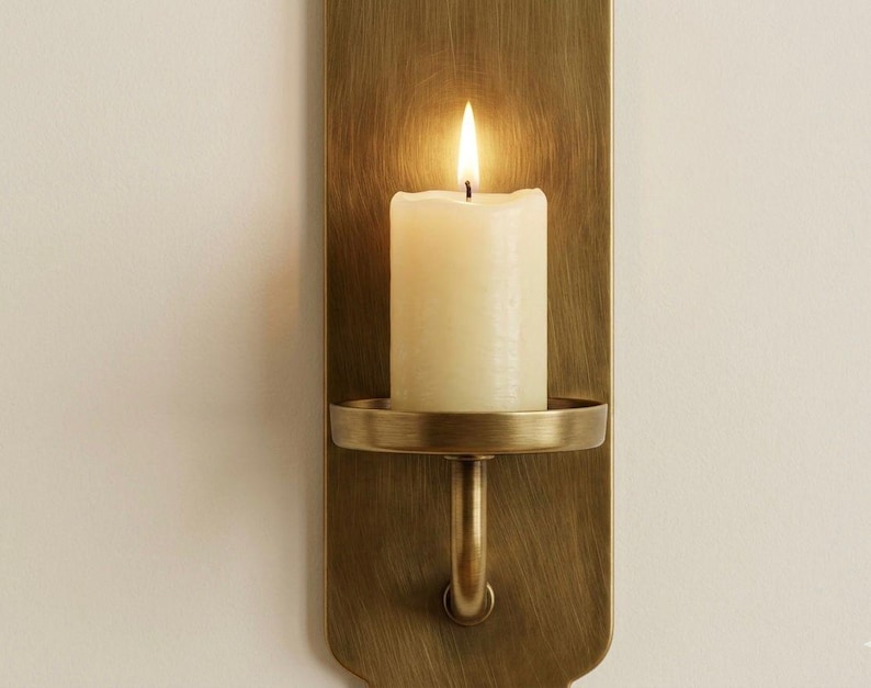 May include: A lit ivory candle sits in a brass wall sconce. The sconce has a rectangular backplate and a circular candle holder. The flame is bright against the warm tones of the brass and candle.
