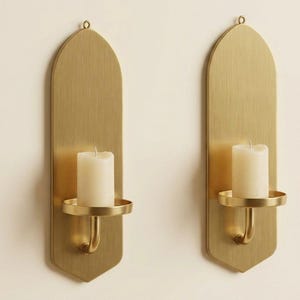 May include: Two brass wall sconces with a vertical, arched design. Each sconce features a round candle holder and a white pillar candle. The sconces are mounted on a light-colored wall, creating a warm, inviting ambiance.