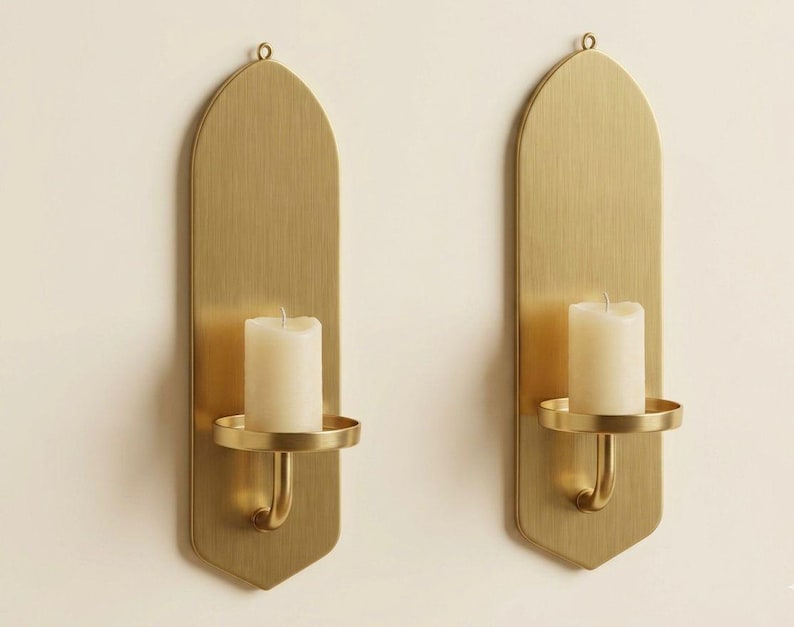 May include: Two brass wall sconces with a vertical, arched design. Each sconce features a round candle holder and a white pillar candle. The sconces are mounted on a light-colored wall, creating a warm, inviting ambiance.