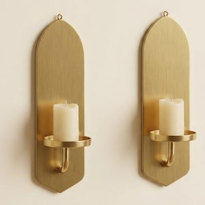 May include: Two brass wall sconces, each with a tall, arched backplate and a circular candle holder. A white pillar candle sits in each holder. The sconces are mounted on a cream-colored wall.