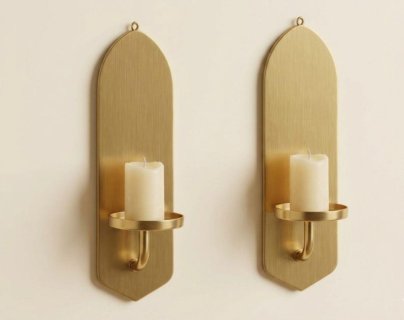 May include: Two brass wall sconces, each with a tall, arched backplate and a circular candle holder. A white pillar candle sits in each holder. The sconces are mounted on a cream-colored wall.