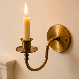 May include: A wall-mounted brass candle holder with a curved arm and a circular backplate secured by two screws. The holder features a central cup with a wide drip tray and contains a single cream-colored taper candle with a lit flame.