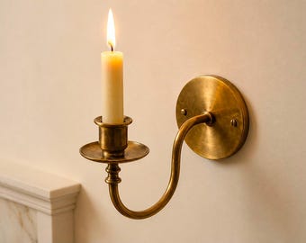May include: A wall-mounted brass candle holder with a curved arm and a circular backplate secured by two screws. The holder features a central cup with a wide drip tray and contains a single cream-colored taper candle with a lit flame.