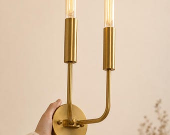 May include: A dual-arm wall sconce featuring a brushed brass finish with two vertical cylindrical sockets. Each socket holds a long, slender, glowing Edison-style tubular light bulb. The fixture is mounted on a circular brass wall plate.