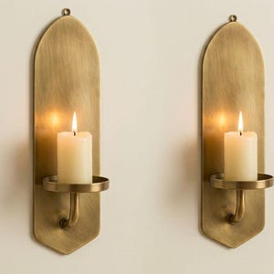 May include: Two brass wall sconces with a warm, antique finish. Each sconce features a long, arched backplate, a circular candle holder, and a lit white pillar candle. The sconces are mounted on a neutral-colored wall, creating a warm and inviting ambiance.