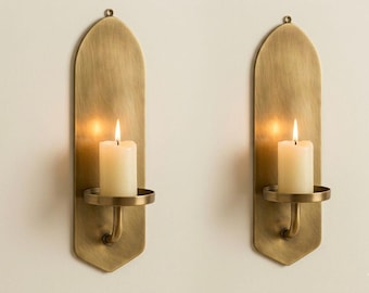 May include: Two brass wall sconces with a warm, antique finish. Each sconce features a long, arched backplate, a circular candle holder, and a lit white pillar candle. The sconces are mounted on a neutral-colored wall, creating a warm and inviting ambiance.