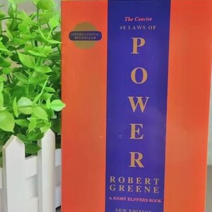 Pu&ograve; includere: Un libro intitolato The Concise 48 Laws of Power di Robert Greene, un libro di Joost Elffers, nuova edizione. La copertina presenta una striscia verticale viola centrata tra due pannelli arancioni, con un sigillo dorato International Bestseller a sinistra.