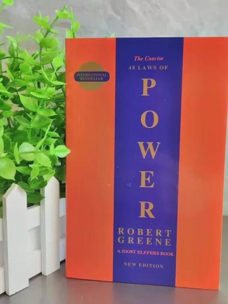Pu&ograve; includere: Un libro intitolato The Concise 48 Laws of Power di Robert Greene, un libro di Joost Elffers, nuova edizione. La copertina presenta una striscia verticale viola centrata tra due pannelli arancioni, con un sigillo dorato International Bestseller a sinistra.