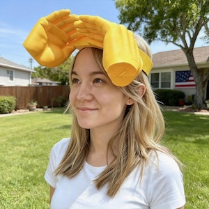 May include: A yellow hand-shaped headband. The headband features two large, open hands positioned above the head, with the palms facing forward. The hands are a bright yellow color and appear to be made of a lightweight material. The headband is worn by a person.