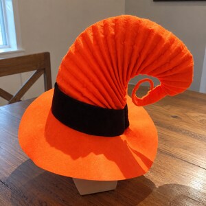 Peut inclure: Un chapeau de sorcière orange vif doté d'un large bord circulaire et d'une haute couronne plissée et incurvée qui se termine par une pointe enroulée. Une bande noire unie entoure la base de la couronne, créant un contraste avec le tissu orange.