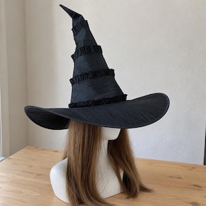 May include: A black pointed hat with a wide brim and a tall, conical crown featuring horizontal rows of gathered fabric ruffles. The hat is displayed on a mannequin head with long, straight, light brown hair, showcasing the textured material.