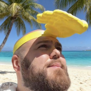 May include: A person wearing a yellow headband and a large yellow hand-shaped hat, looking upwards. The background shows a tropical beach with palm trees, white sand, and turquoise water under a clear blue sky.
