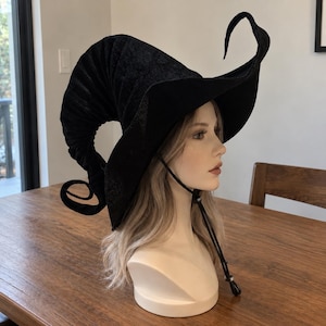 Peut inclure: Un chapeau de sorcière en velours noir avec une couronne haute, courbée et pointue, doté d'un large bord ondulé. Le chapeau comprend une fine mentonnière noire avec un cordon de serrage réglable, présenté sur un mannequin à cheveux clairs.