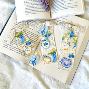 Puede incluir: Tres marcapáginas rectangulares transparentes con ilustraciones coloridas de bayas azules, tulipanes amarillos, flores blancas, patos y sombreros azules. Cada marcapáginas está rematado con una cinta de encaje color crema y un pequeño dije.
