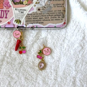 May include: Two gold-tone metal charms attached to clear plastic clips. One charm features a pink circle with the letter J, a red chilli pepper, and red cherries. The other charm features a pink circle with the letter M, red cherries, and a pink oval gem.