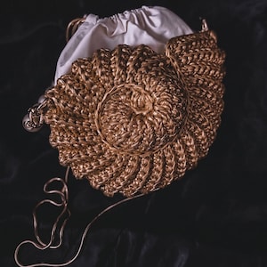 Può includere: Una borsa a mano dorata intrecciata con un design a conchiglia a spirale. La borsa ha un sacchetto bianco con coulisse all'interno e una delicata tracolla a catena dorata. La borsa è su uno sfondo scuro.